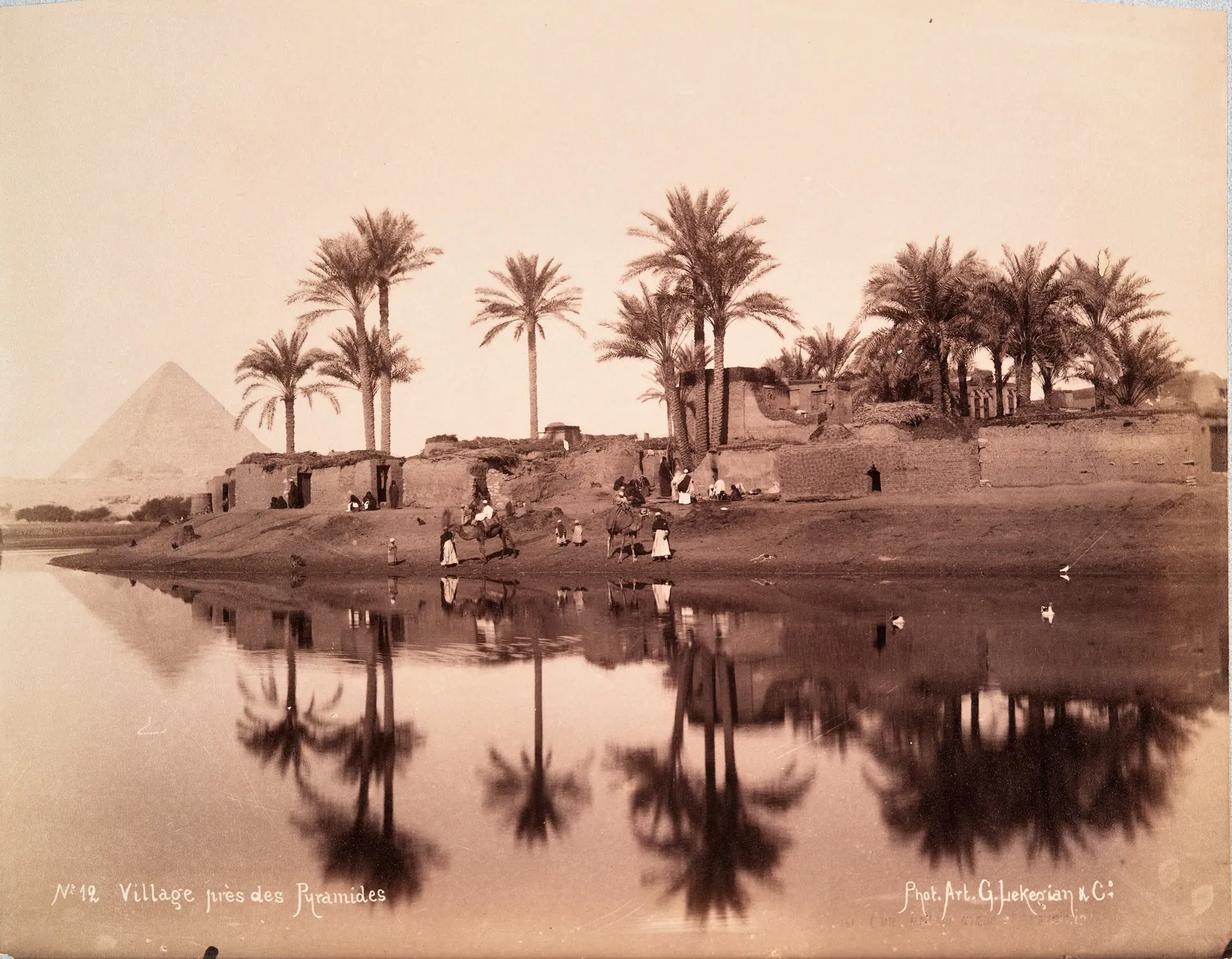 ”Village près des Pyramides” – G. Lekegian, ca 1880-tal: lerhus och palmer speglas i översvämningsvattnet, med en pyramid vid horisonten. Fram till Assuanhöga dammen nådde den årliga översvämningen nästan ända fram till platåns fot varje höst – i mitten av oktober var den nära sin höjdpunkt.