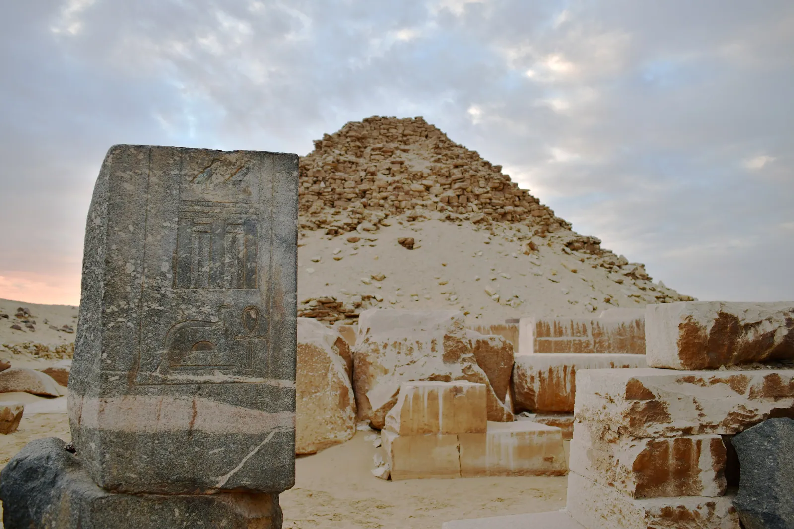 Ett mörkt granitblock med ristat palatsfasadmotiv (serekh/skendörr) framför Sahures pyramid – en påminnelse om de dyrbara hårda bergarter som användes i templet.