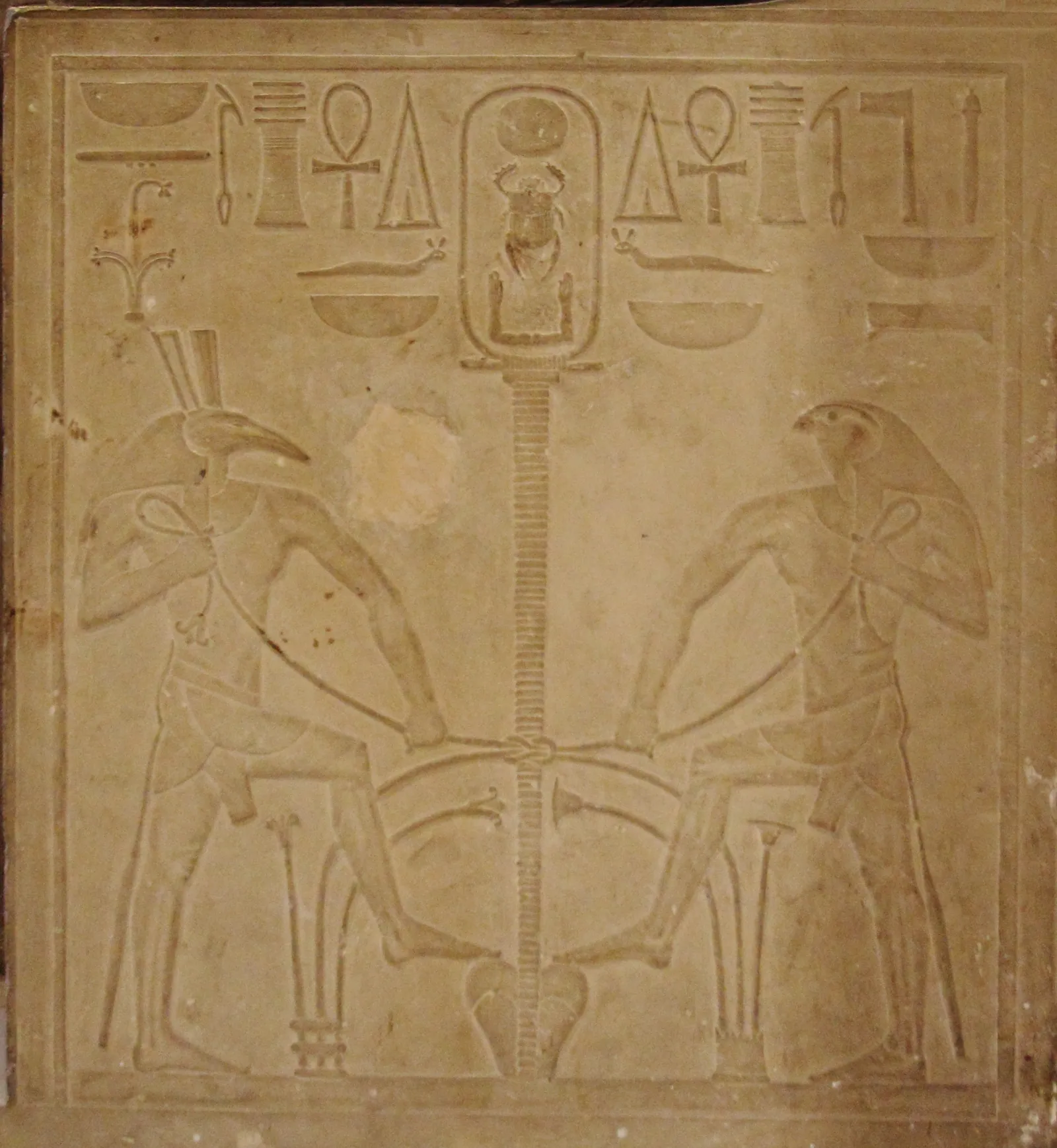 Tronsidan på en av de tio statyerna från Lisht: Horus och Seth knyter samman Över- och Underegyptens växter kring hieroglyfen för ”förena” (sema-tawy). På fem av statyerna är det de två gudarna, på de övriga fem två nilgudar (Hapi). Jämför dem på GEM.